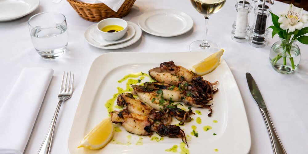 Assiette de petits calmants ou chipirons grillés nappés d'une persillade et accompagnés de citron, dressée sur une table nappée de blanc au restaurant El Faro dans le quartier des Pâquis à Genève