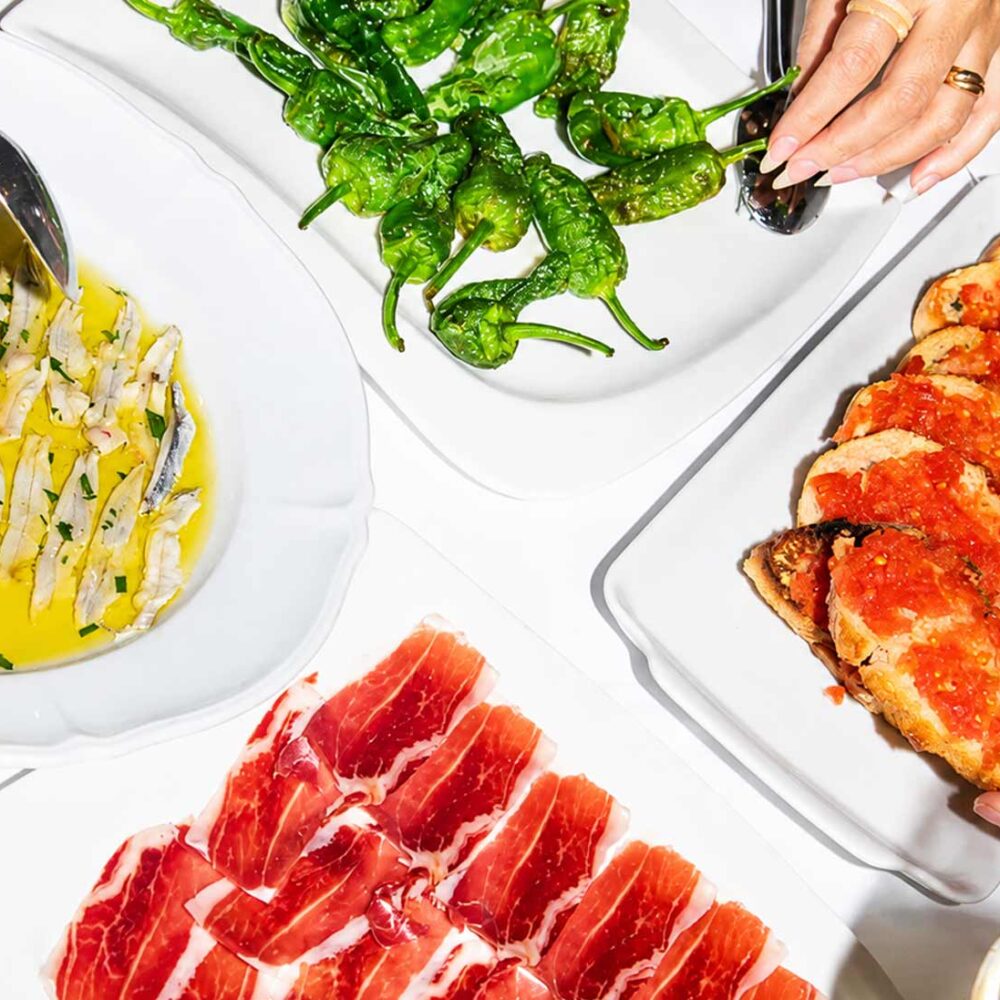 Table garnie de plusieurs assiettes de tapas espagnoles comprenant du jambon ibérique, des pimientos de Padrón, du pan con tomate et des anchois, servie au restaurant El Faro dans le quartier des Pâquis à Genève